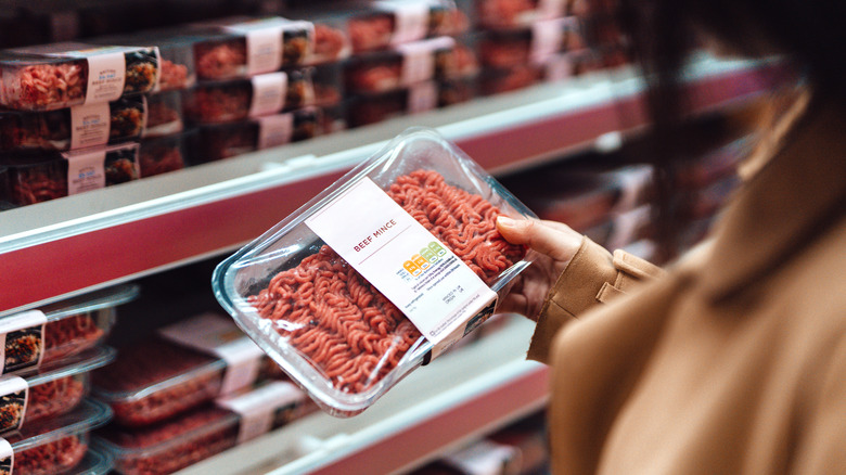 Above the shoulder view of woman holding a pack of beef mince in a supermarket. Standing in front of the meat product aisle and have no idea which product to choose from. The Conscious Carnivore - find meat that is locally-sourced, healthy, high quality butchering, and handled with respect. The environmentally conscious way to choosing food.